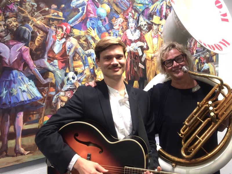 Julius mit Gitarre und Heike Besen am Sousaphon vor einem farbenfrohen Gemälde von Werner Petzold.
